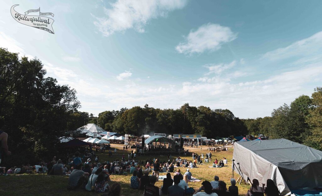 Ein weitläufiges Festivalgelände im Freien, auf dem zahlreiche Menschen auf einer Wiese sitzen und einer Hauptbühne entgegenblicken. Mehrere Zelte und Stände sind im Hintergrund zu sehen, umgeben von Bäumen. Der Himmel ist teils bewölkt, und es herrscht eine entspannte Atmosphäre